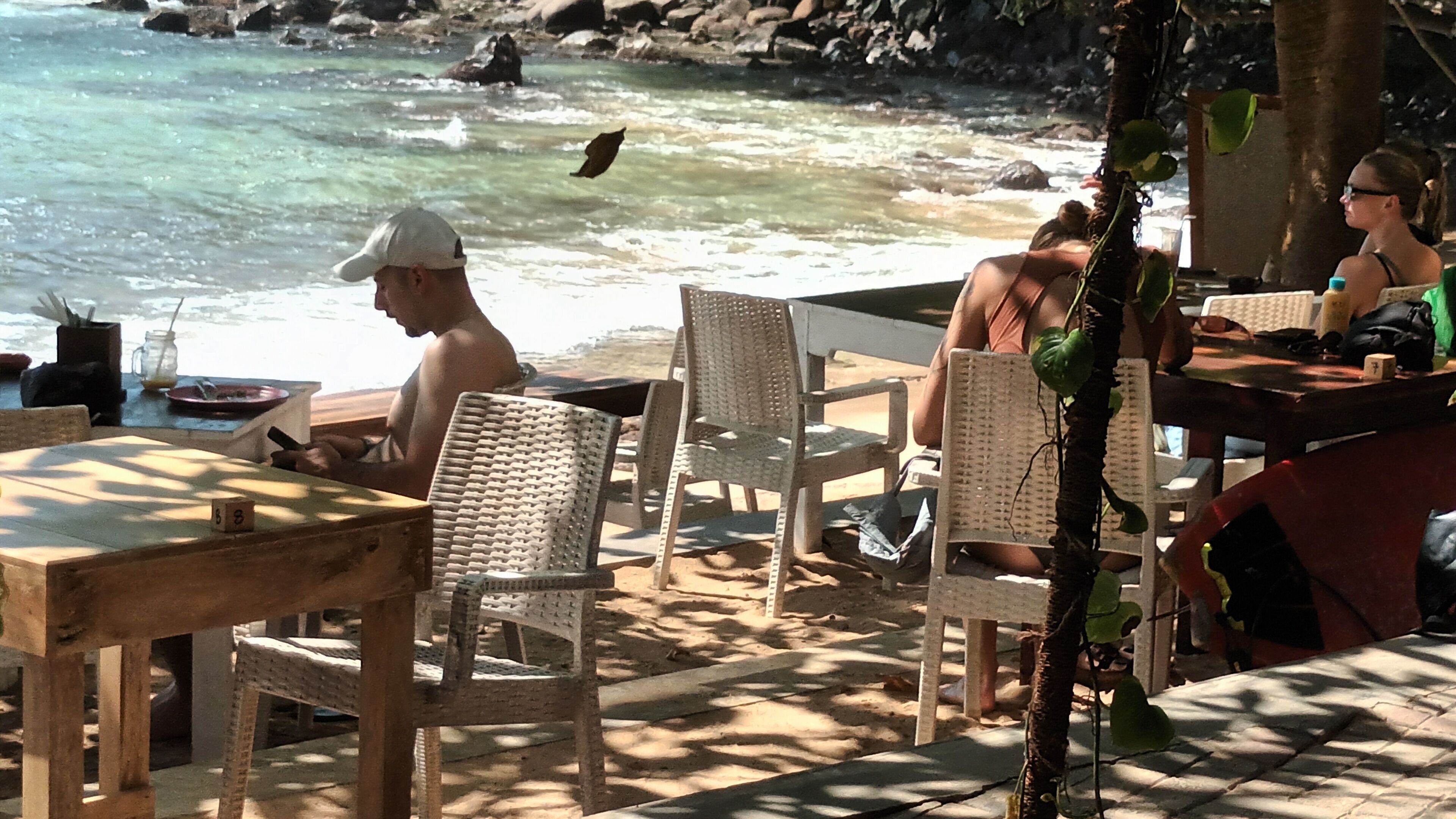 An outdoor seating area with wicker chairs and tables, overlooking a beach with clear water and rocks.