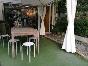 Een terras met een houten tafel en witte stoelen, omgeven door groen en een glazen wand met artistieke tekeningen.