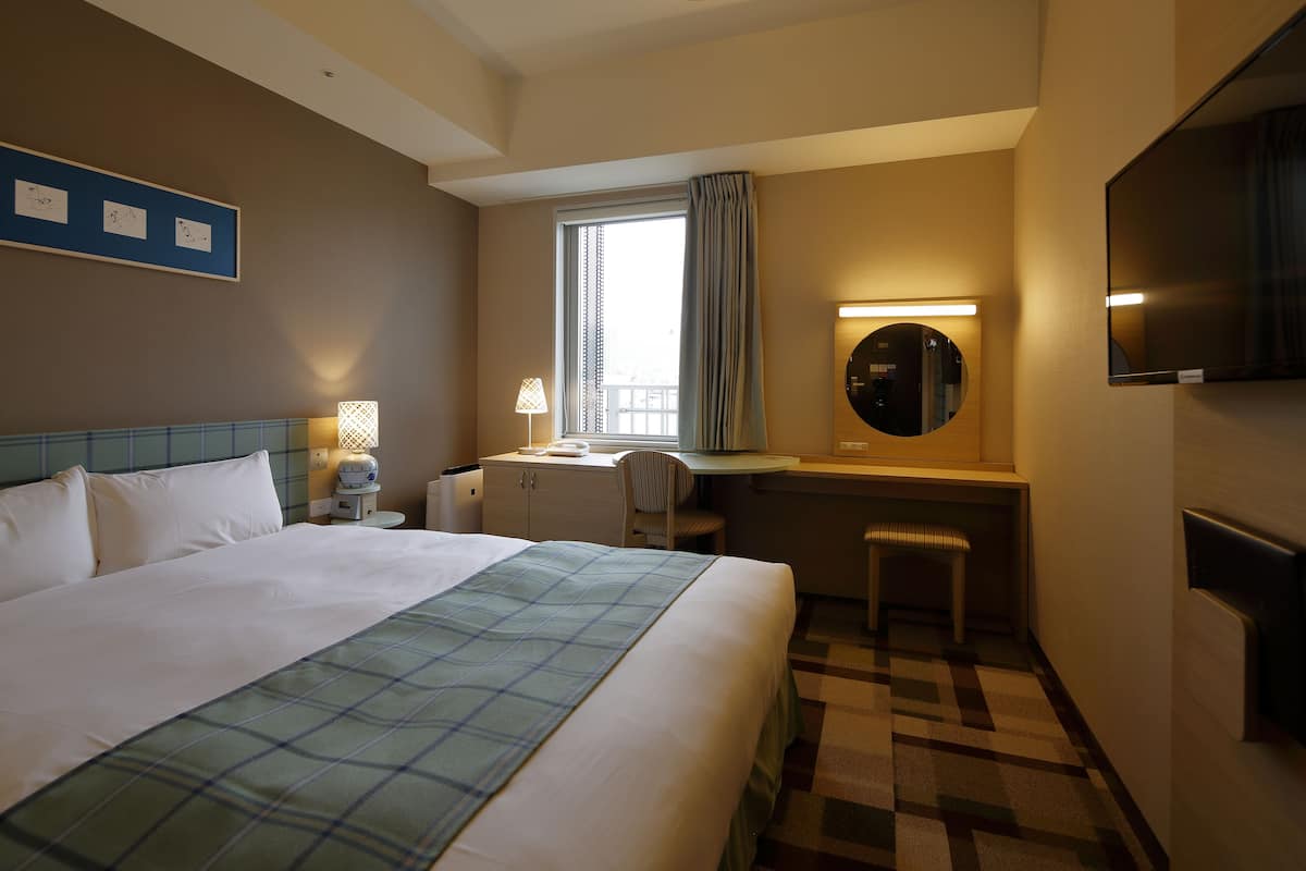 Une chambre d’hôtel avec un grand lit, un bureau avec une chaise, une télévision et une fenêtre avec des rideaux.
