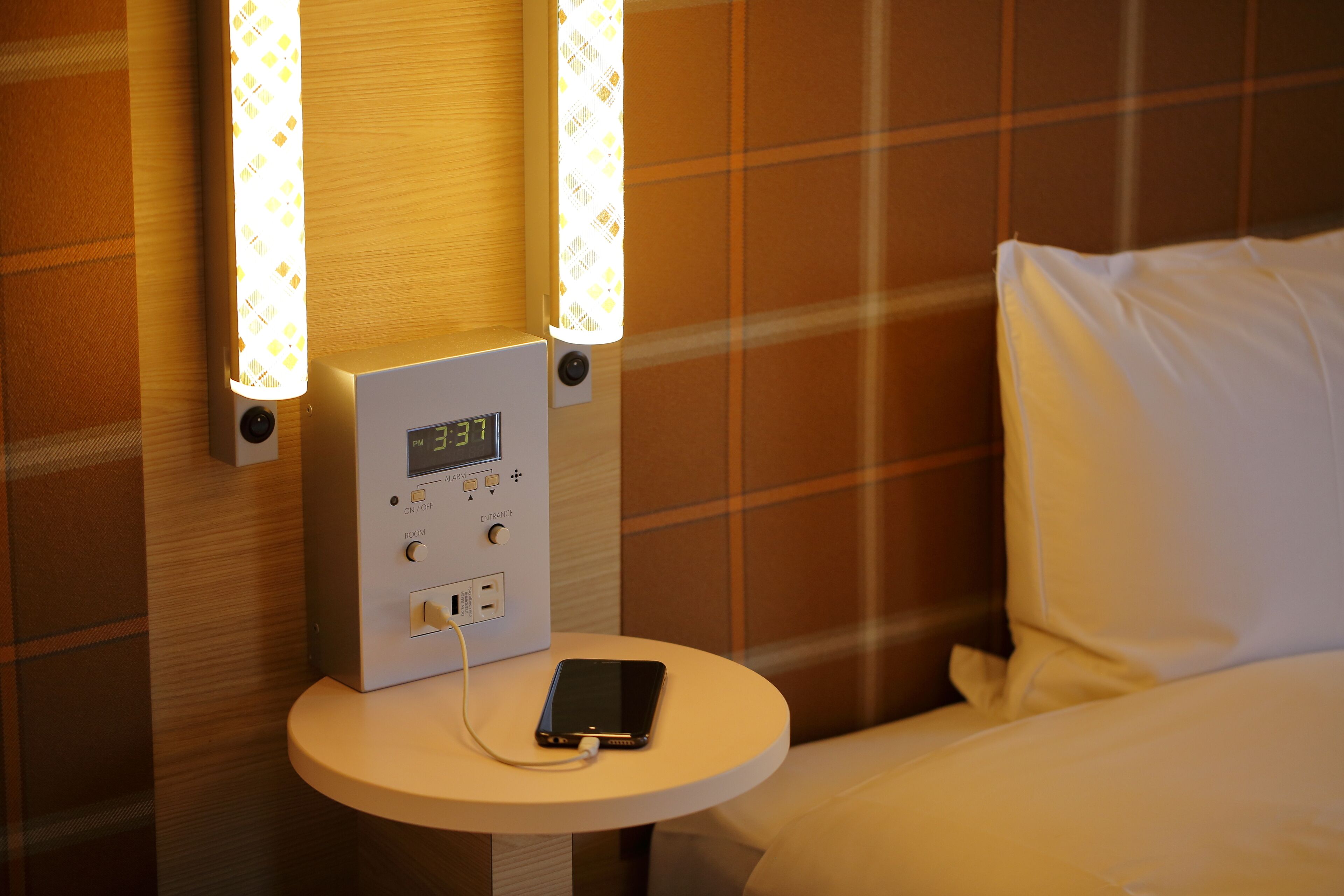 A hotel room with a bedside table, a digital clock, and a smartphone charging.