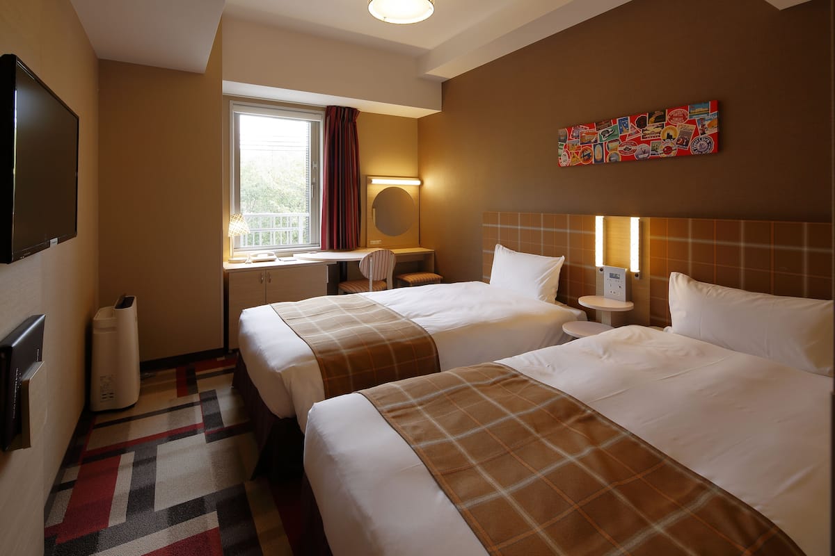 Une chambre d’hôtel avec deux lits, un bureau, une chaise, une télévision, une fenêtre avec des rideaux, un tableau décoratif au mur et une poubelle.