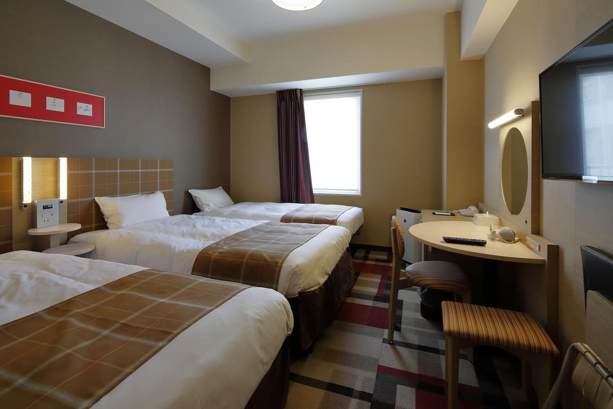 Une chambre d’hôtel avec trois lits, un bureau, une chaise, une télévision et une fenêtre avec des rideaux.