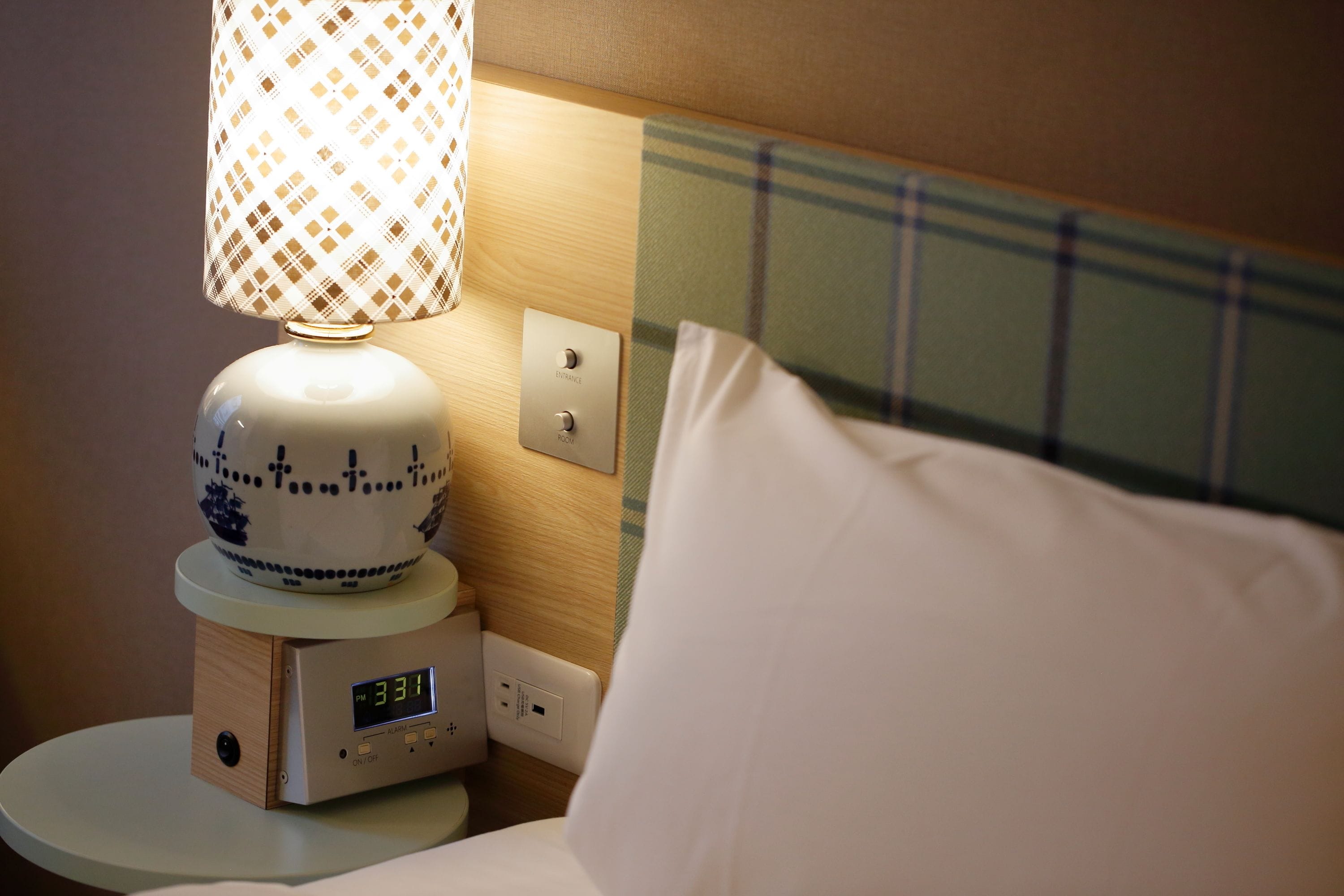 A bedside table with a digital clock, a patterned lamp, and a pillow.