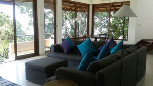 Una sala de estar moderna con un sofá gris, almohadones a rayas azules y rojos, y una mesa de centro redonda. Amplias ventanas ofrecen vistas al exterior.