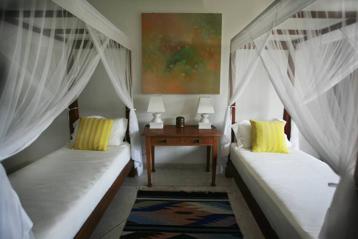 A room with two beds, each with white bedding and yellow pillows, a small table with lamps, and a painting on the wall.