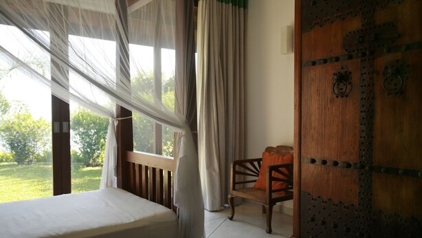 A bedroom with a bed, a chair, a wooden door, and a window with curtains. - Sea Heart House on Koggala Lake (Koggala)