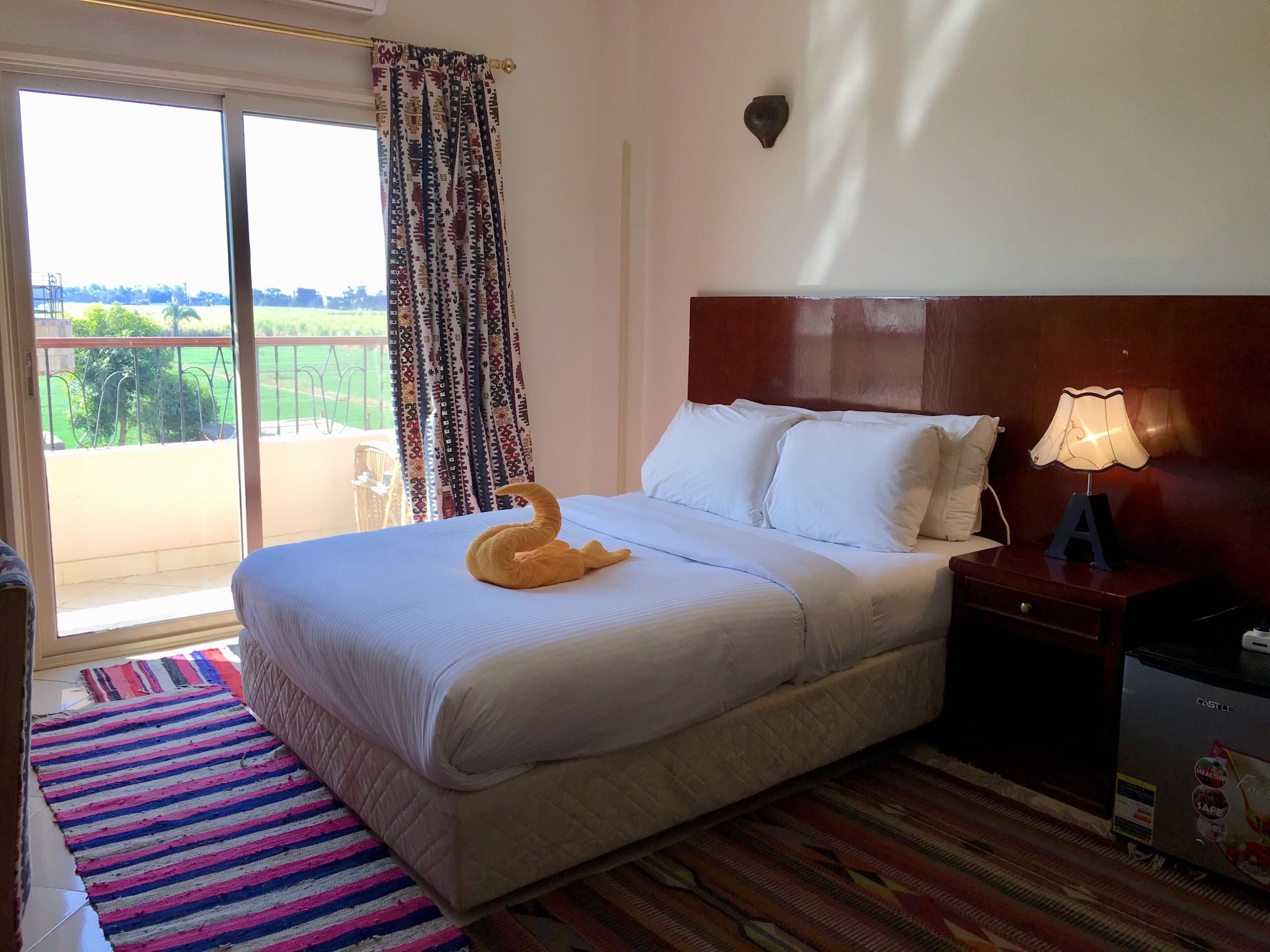 Habitación de hotel con cama, mesita de noche con lámpara, alfombra a rayas y vistas a un balcón con cortina estampada.