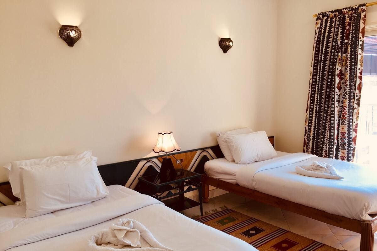 A hotel room with two beds, a nightstand with a lamp, patterned curtains, and a decorative rug.