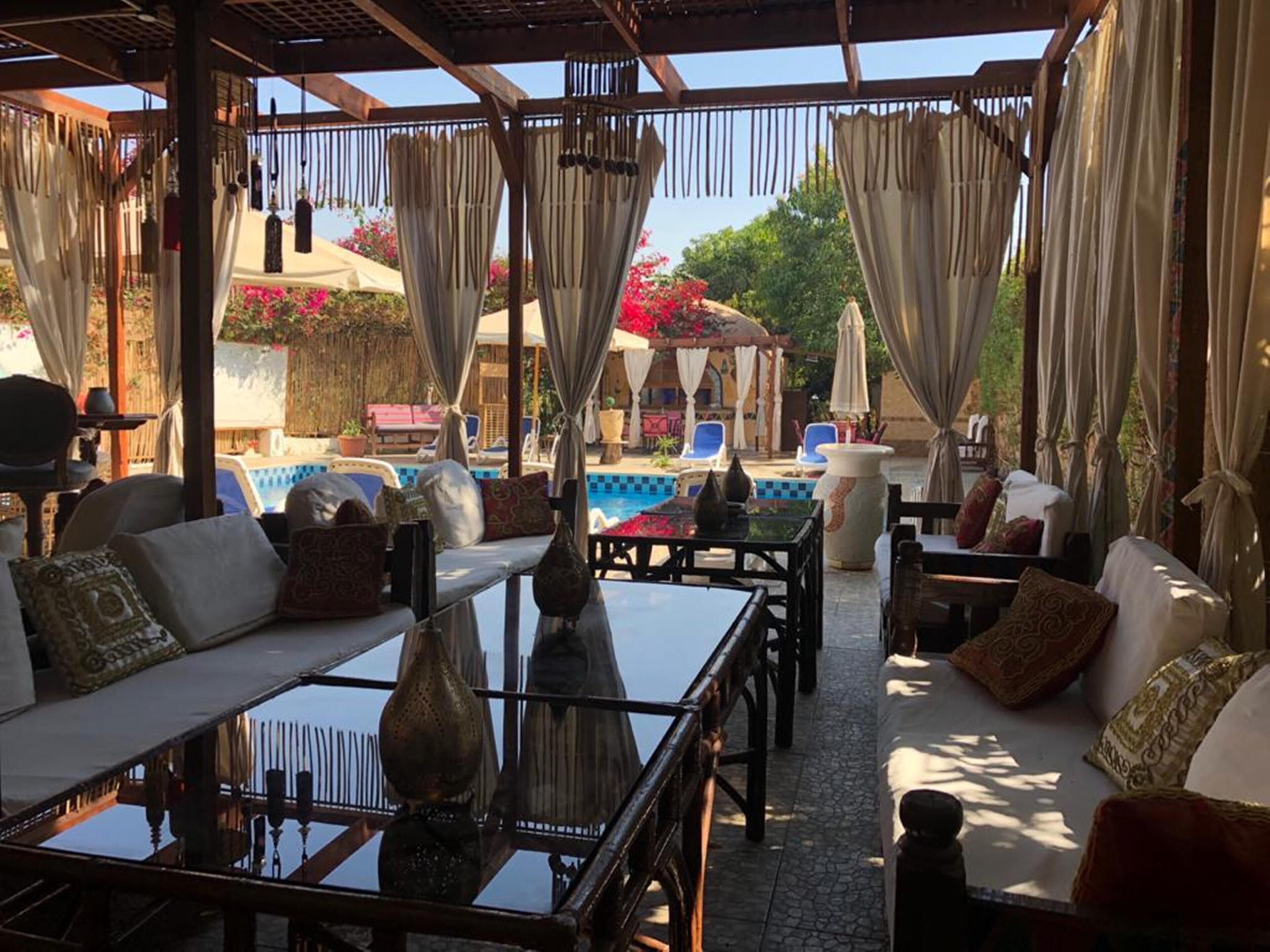 An outdoor seating area with tables and sofas, a pool, and a pergola with curtains.