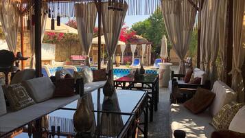 An outdoor seating area with tables and sofas, a pool, and a pergola with curtains.