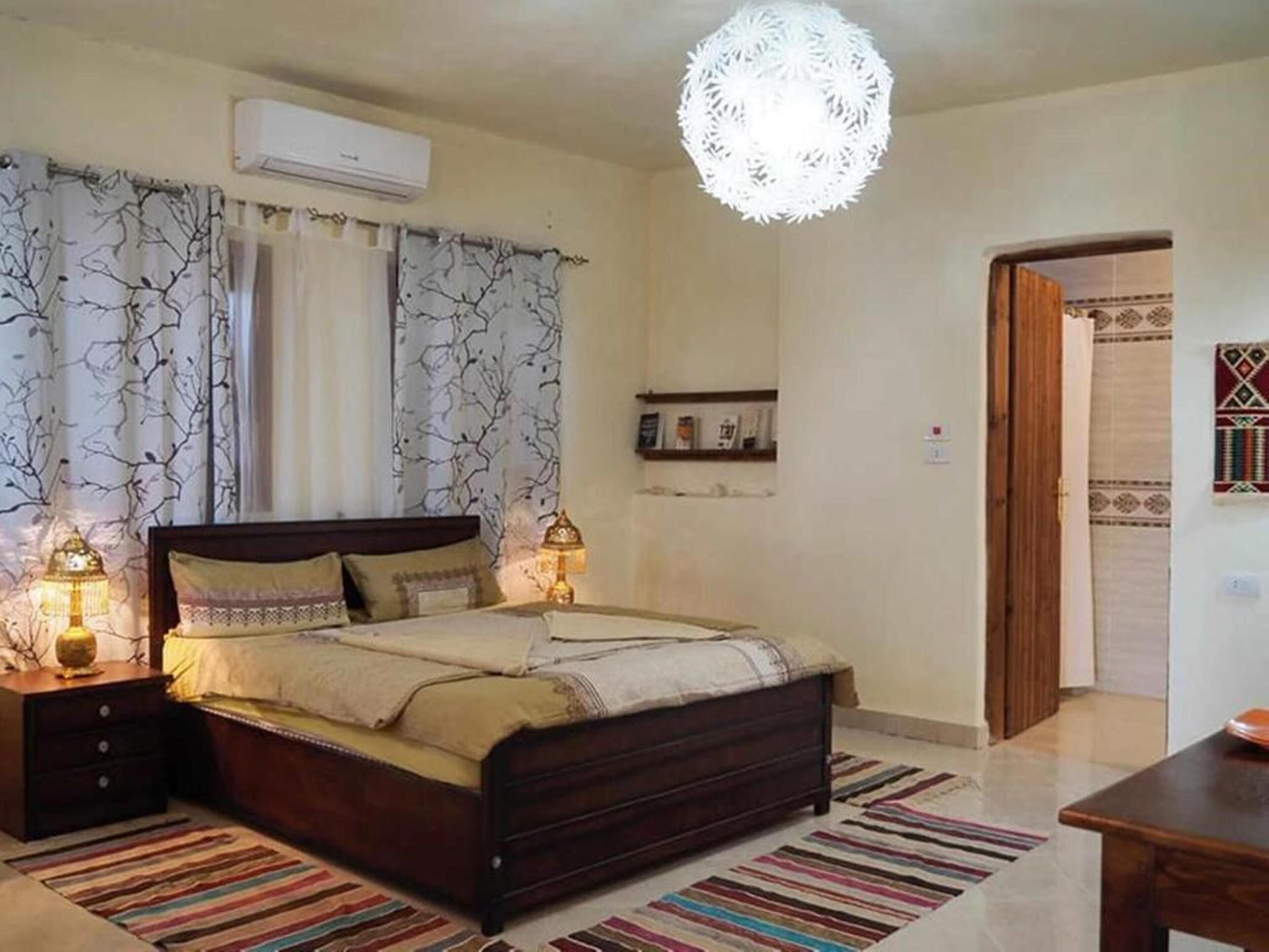A bedroom with a wooden bed, two bedside lamps, a hanging light fixture, patterned curtains, and a small shelf with books.