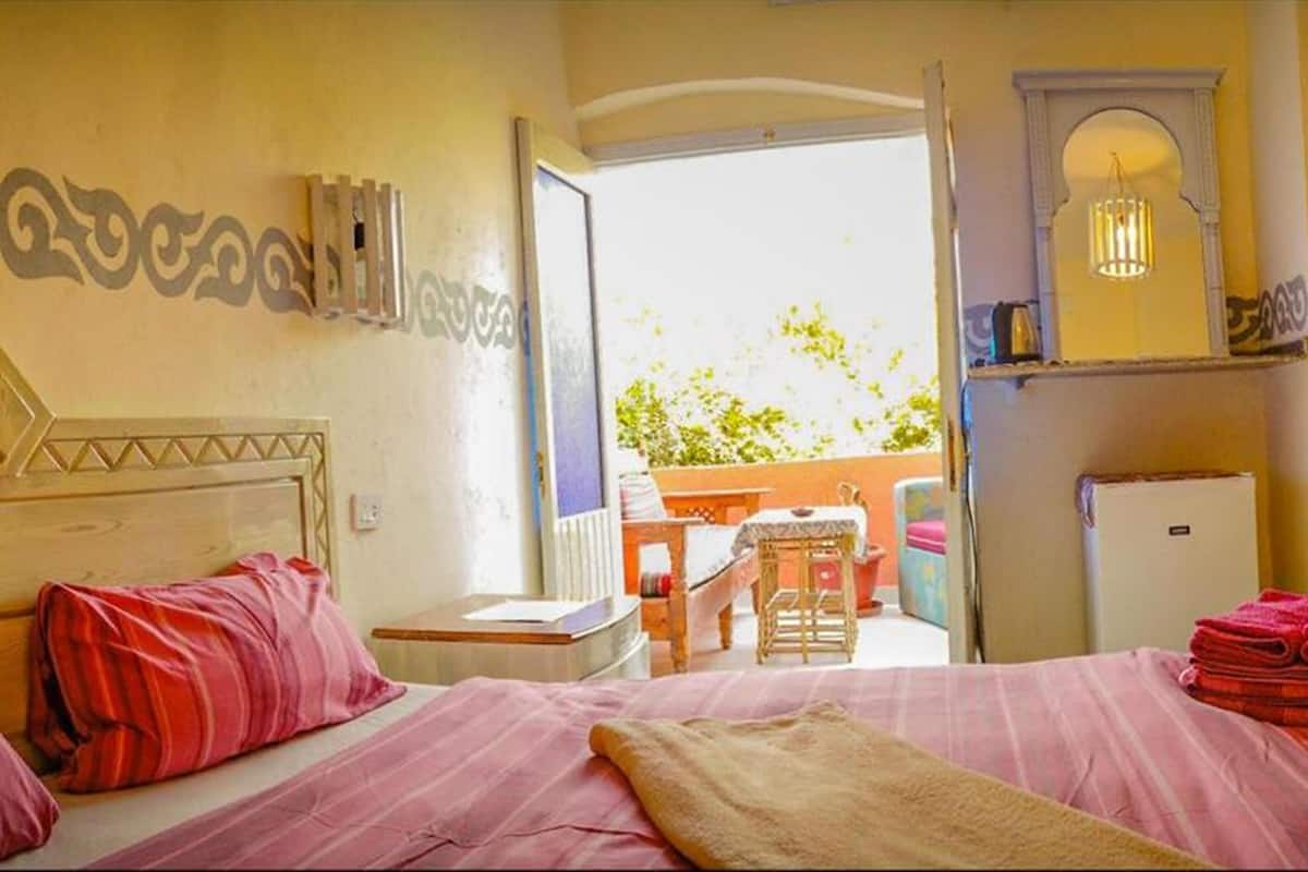 A hotel room with two beds, a small table, a chair, a refrigerator, and a balcony with a view of greenery.