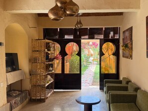 A room with a large open door leading to a garden, a wicker shelf with decorative items, a round table, and green upholstered seating. - Nile Compound Hotel (Luxor)