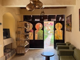 A room with a large open door leading to a garden, a wicker shelf with decorative items, a round table, and green upholstered seating.