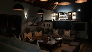 A dimly lit lounge area with multiple seating options, a bar with shelves of bottles, and a rustic ceiling.