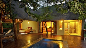 A wooden deck with a pool, a thatched-roof building, and a glass door leading to a bedroom.