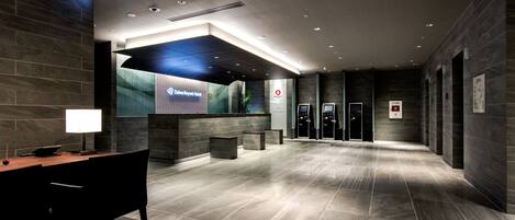 A modern hotel lobby with a reception desk, seating area, and elevators.