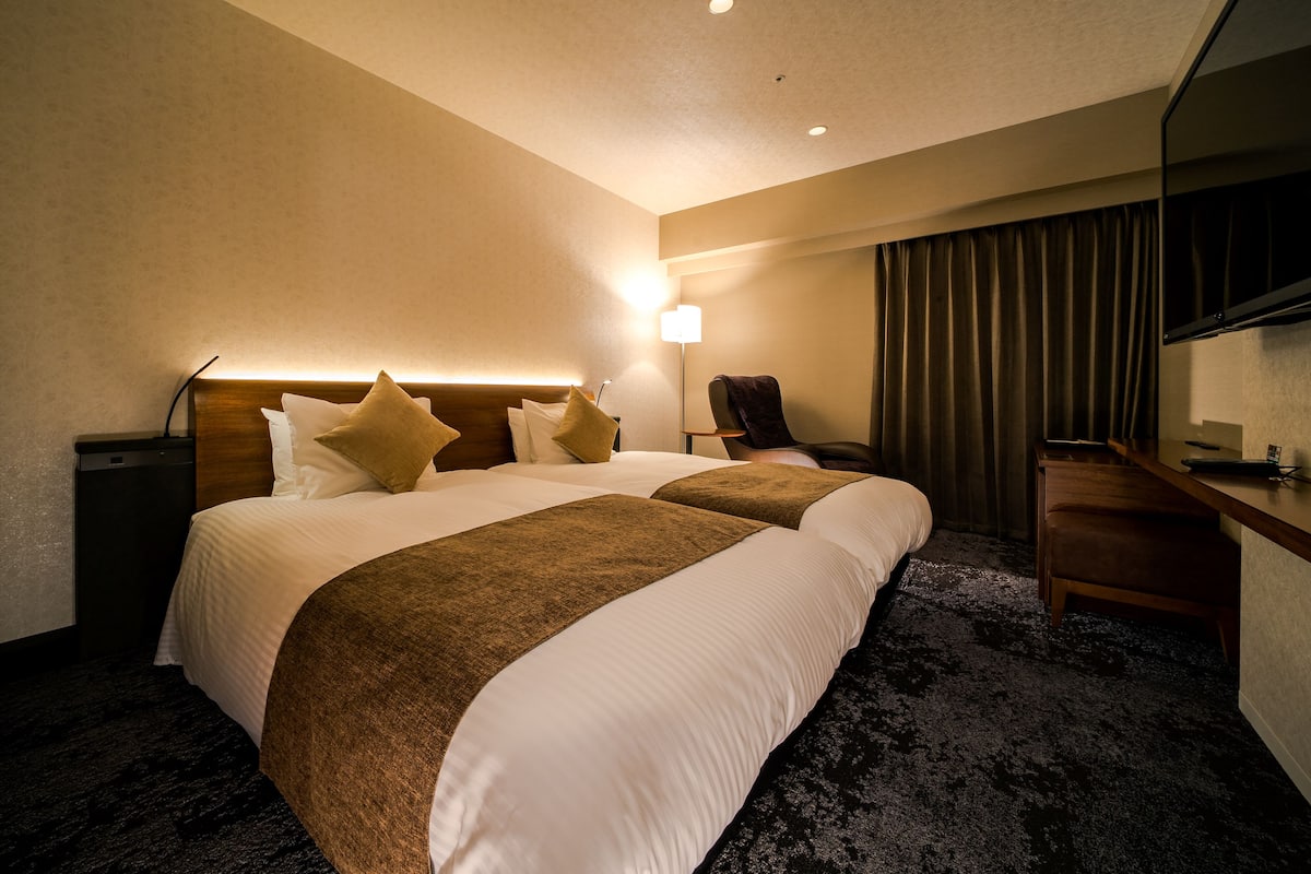 Une chambre d’hôtel avec deux lits, un bureau, une chaise, une lampe et une grande fenêtre avec des rideaux.