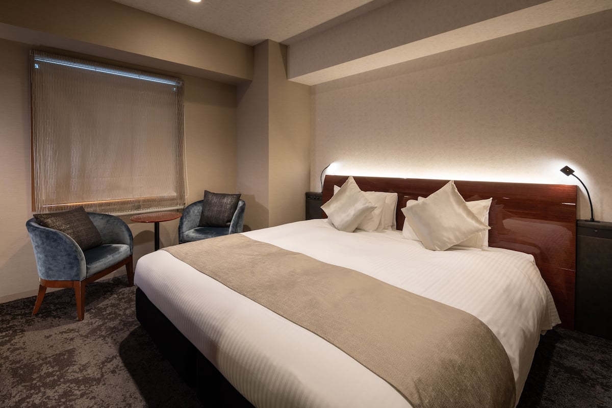 Une chambre d’hôtel avec un grand lit, une table de chevet, une chaise, une fenêtre avec des stores et une lampe.
