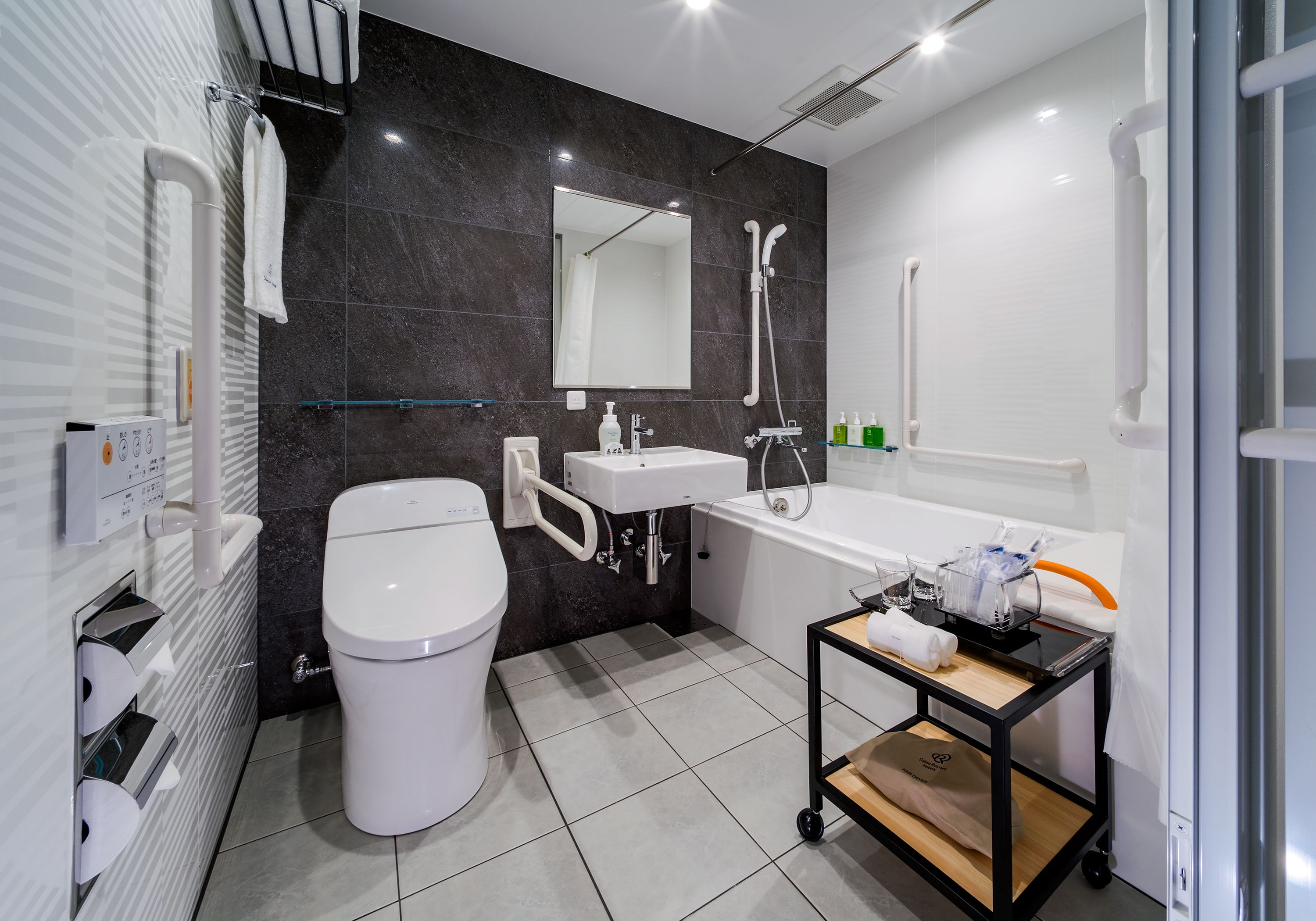Un baño moderno con inodoro, lavamanos y bañera. Hay un pasamanos, un espejo y una mesita de noche con artículos de aseo.