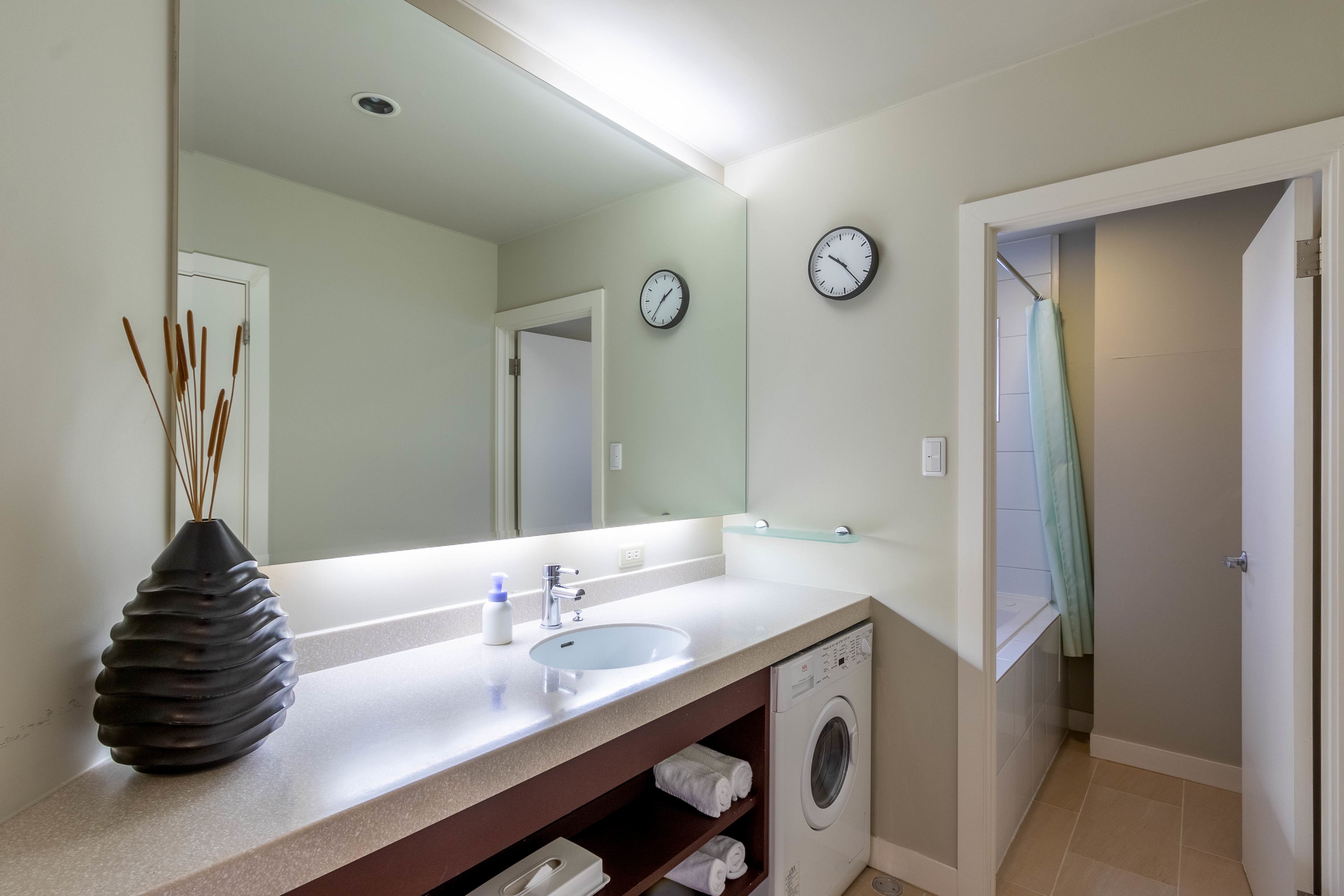 A modern bathroom with a sink, mirror, clock, and a washing machine.