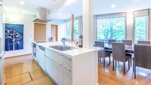 A modern kitchen with a central island, stainless steel appliances, and a dining area with a large window. - Furano Fresh Powder Apartments and Chalet (Furano)