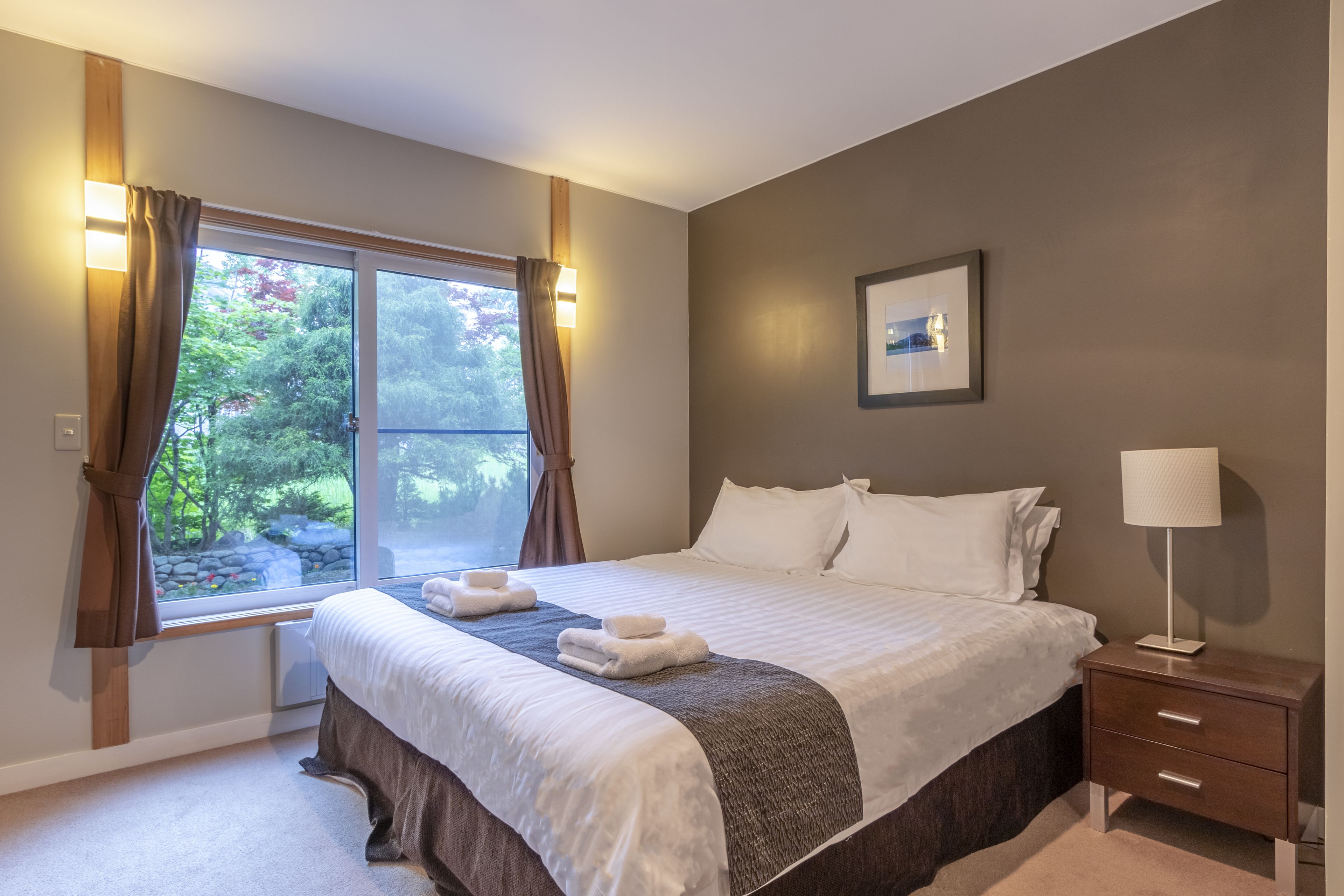 A neatly made bed with white linens, a bedside table with a lamp, and a window with curtains.