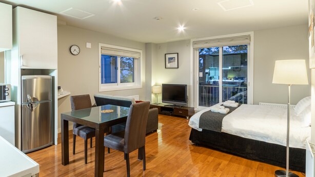 A modern hotel room with a wooden floor, a dining table with chairs, a bed with a white comforter, a TV, and a balcony with a view. - Furano Fresh Powder Apartments and Chalet (Furano)