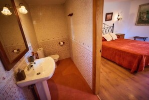 A bathroom with a sink, toilet, and shower. There is a bed with a red blanket in the adjacent room. - As Chivas (Redondela)