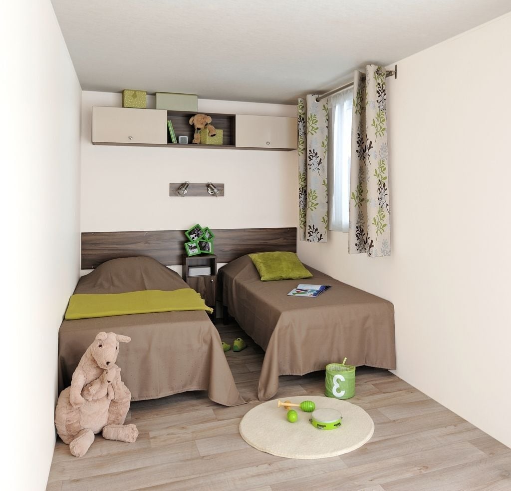 Quarto pequeno e bem arrumado, com duas camas, uma estante com objetos decorativos, um urso de pelúcia e um tapete redondo.