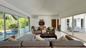 Una sala de estar moderna con un sofá grande, una mesa de centro y un televisor. Hay ventanas amplias y vistas a zonas verdes.
