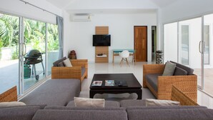 Un salón moderno con un sofá grande, una mesa de centro y un televisor. Hay un balcón con una parrilla y vistas a palmeras.