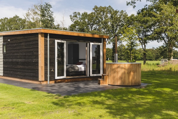 A modern wooden cabin with a glass door entrance, a small wooden deck, and a hot tub in a grassy outdoor area.
