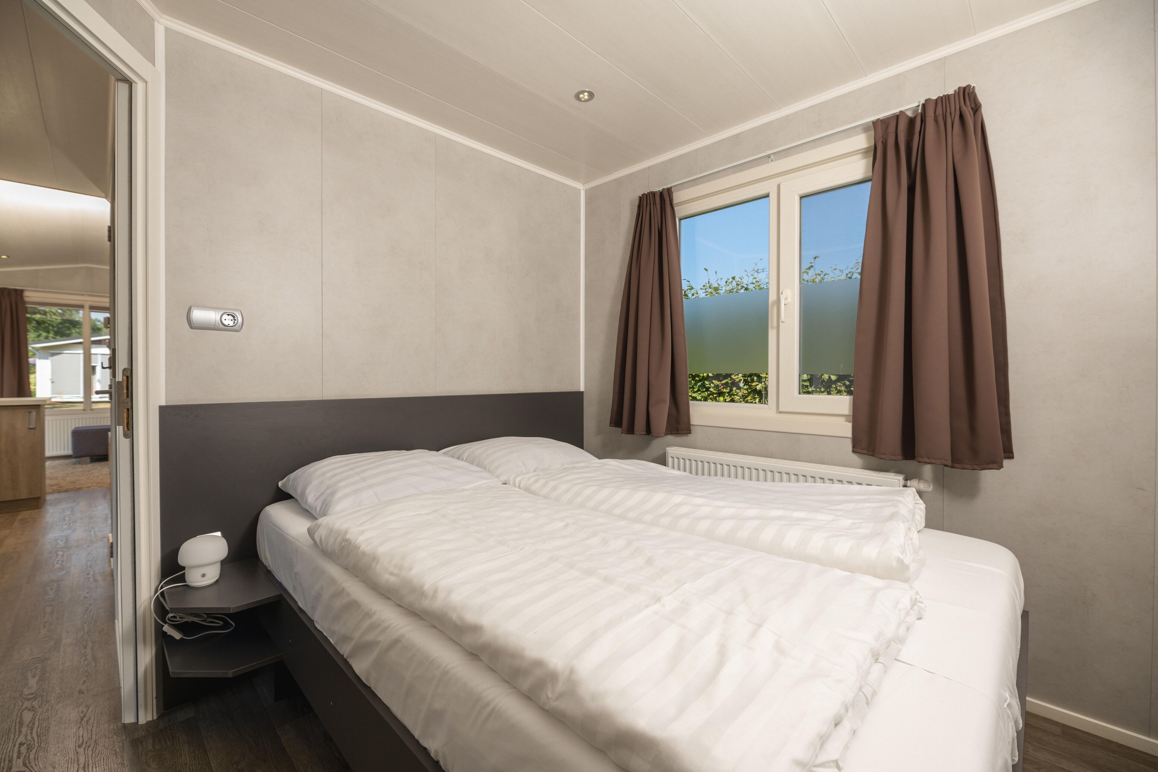 Een slaapkamer met twee bedden, een raam met gordijnen, een nachtkastje met een lamp en een deur naar een andere kamer.