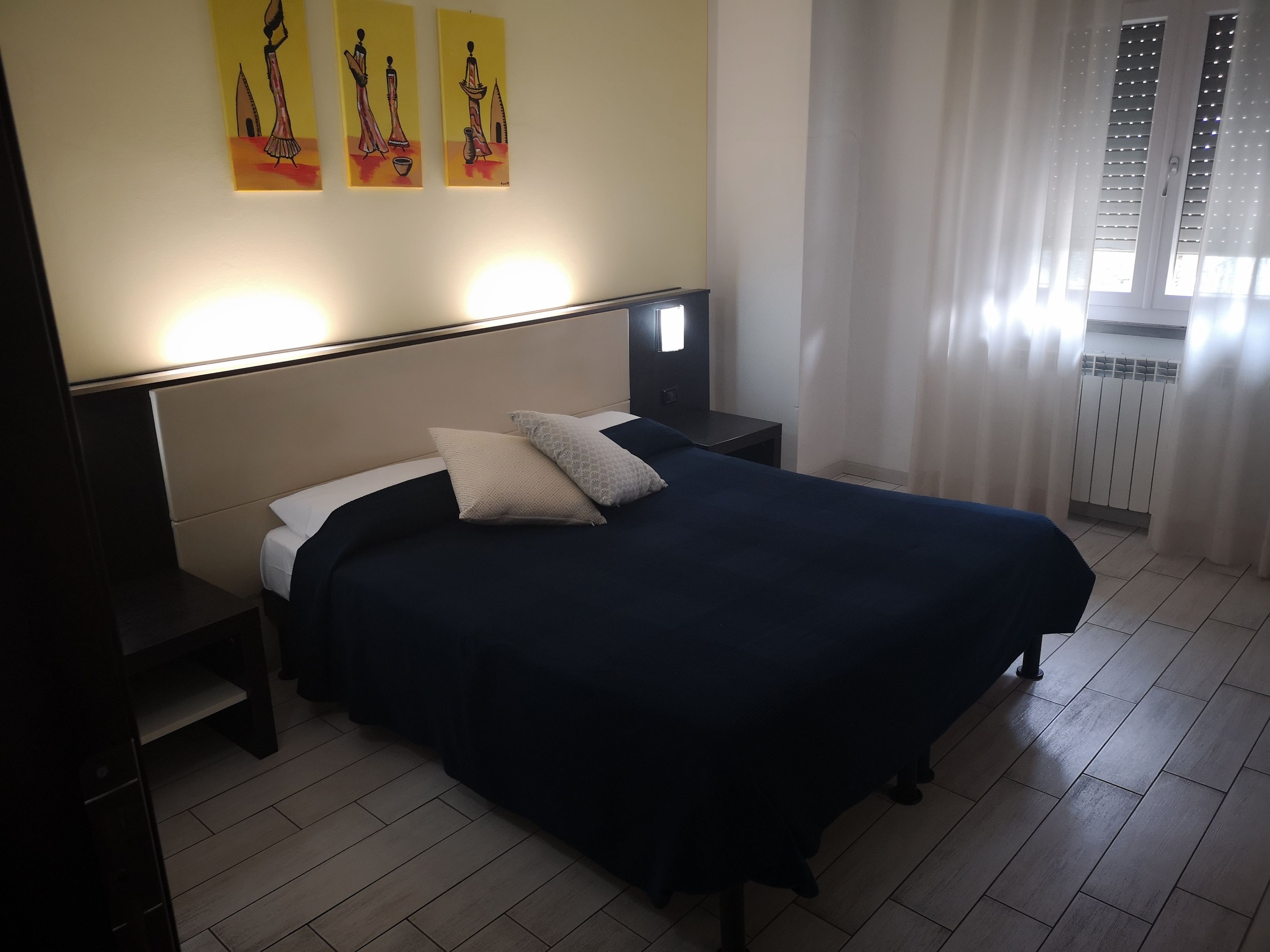 Een keurig opgemaakt bed met een donkerblauwe sprei en twee kussens, een nachtkastje met een lamp, en een muur met drie ingelijste kunstwerken.