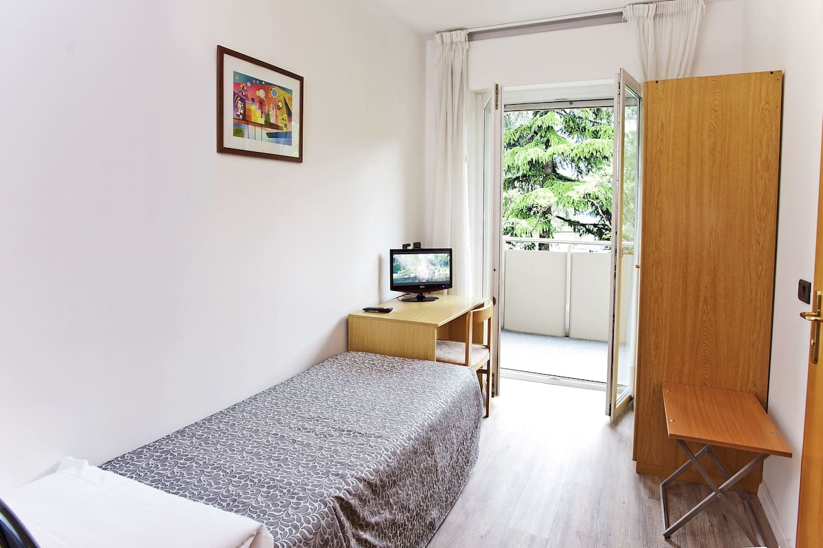 A small, tidy room with a bed, a desk with a computer, a wardrobe, and a door leading to a balcony.