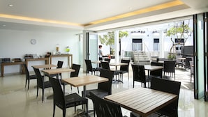 Un café moderno con mesas de madera y sillas de mimbre negro, una barra con un reloj y vistas a un edificio.