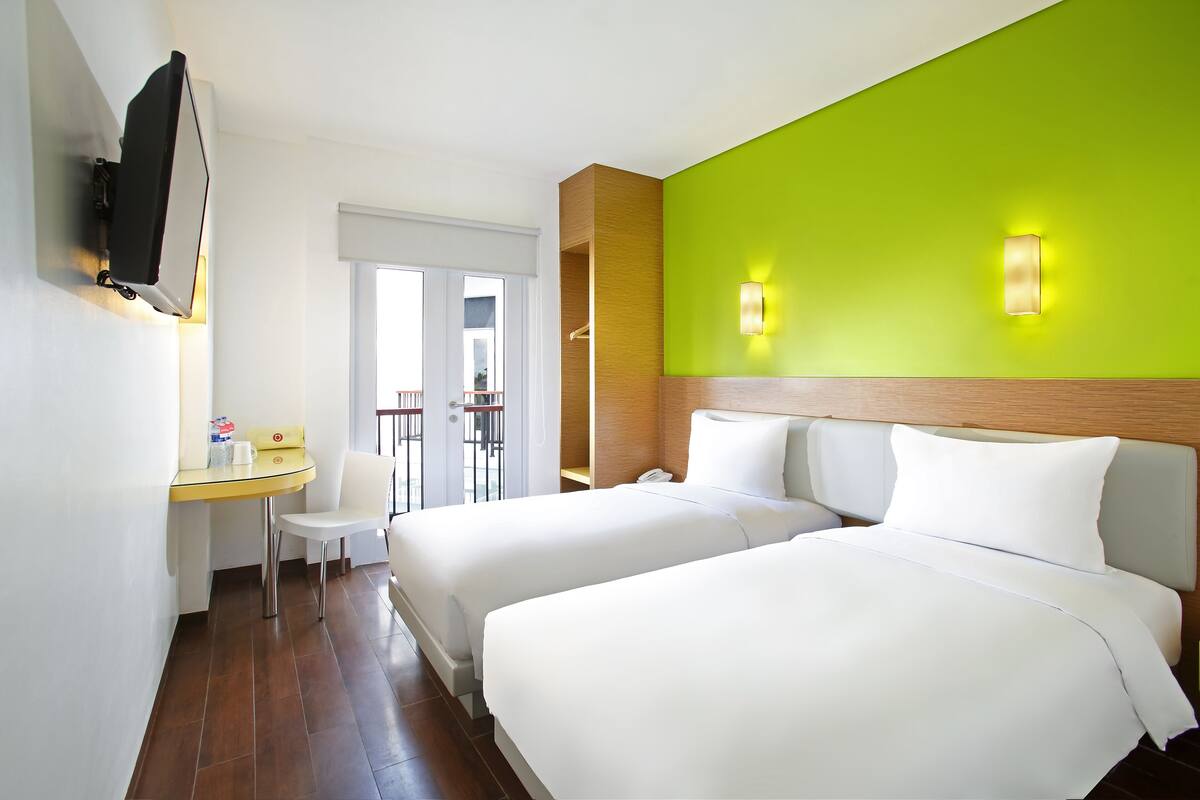 A hotel room with two beds, a small table with a chair, a TV mounted on the wall, and a bright green accent wall.