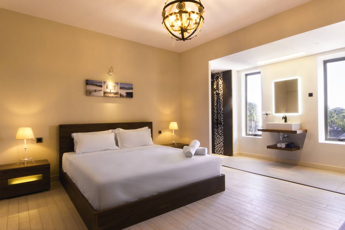 Une chambre d’hôtel moderne dotée d’un grand lit, de lampes de chevet, d’une œuvre d’art accrochée au mur, d’une salle de bain avec un meuble-lavabo et un miroir, et de grandes fenêtres.