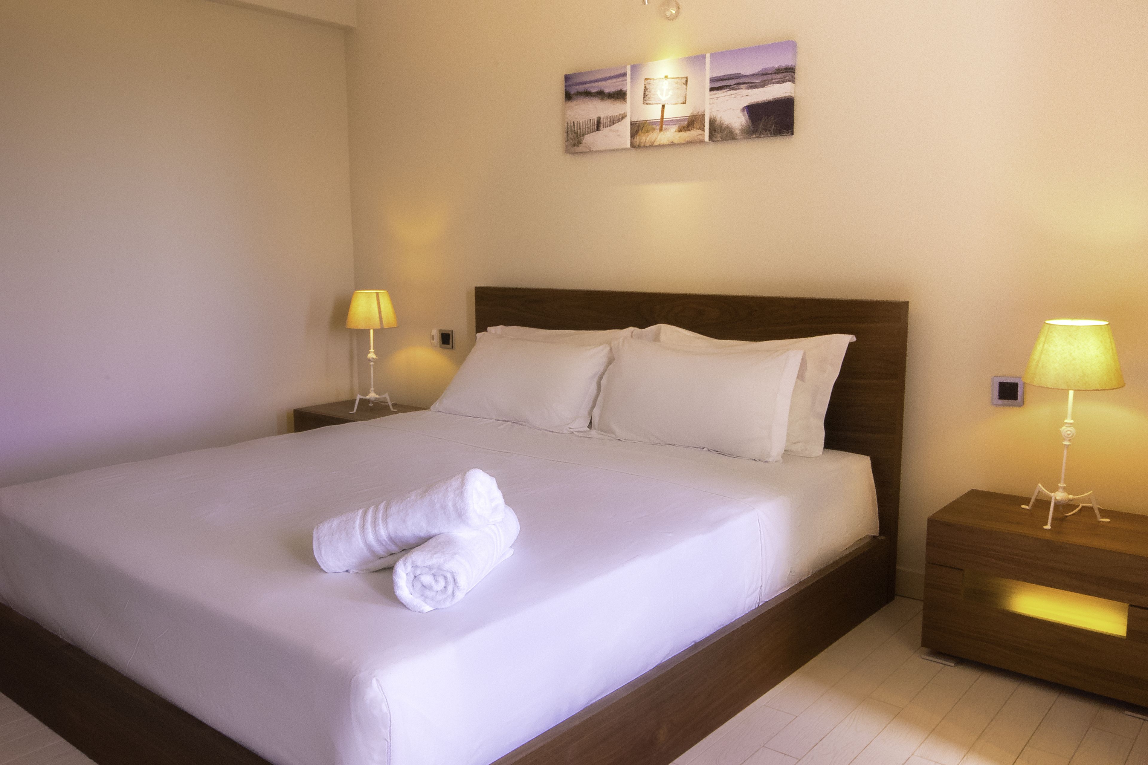 A neatly made bed with white linens and a rolled towel, two bedside lamps, and a wall-mounted picture.