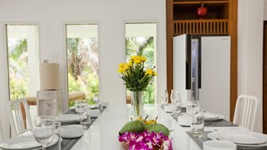 Una mesa de comedor dispuesta con platos blancos, vasos y cubiertos, un florero con flores amarillas y un refrigerador al fondo.