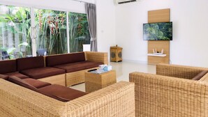 Una sala de estar moderna con un sofá marrón, sillas de mimbre, un televisor de pantalla plana y un ventanal con vistas a la vegetación.