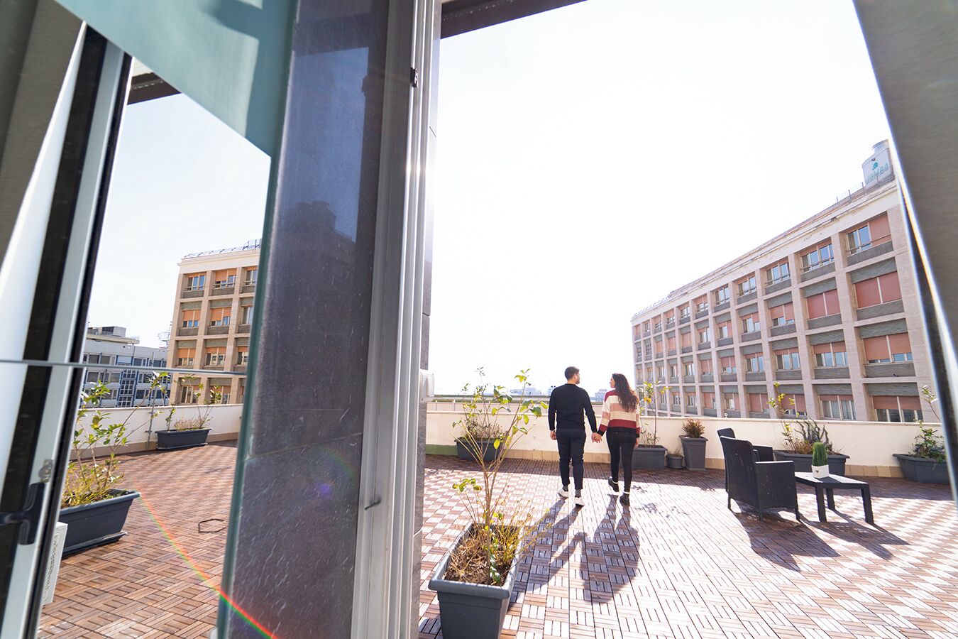 A rooftop terrace with potted plants, a brick floor, and a view of a multi-story building in the background.