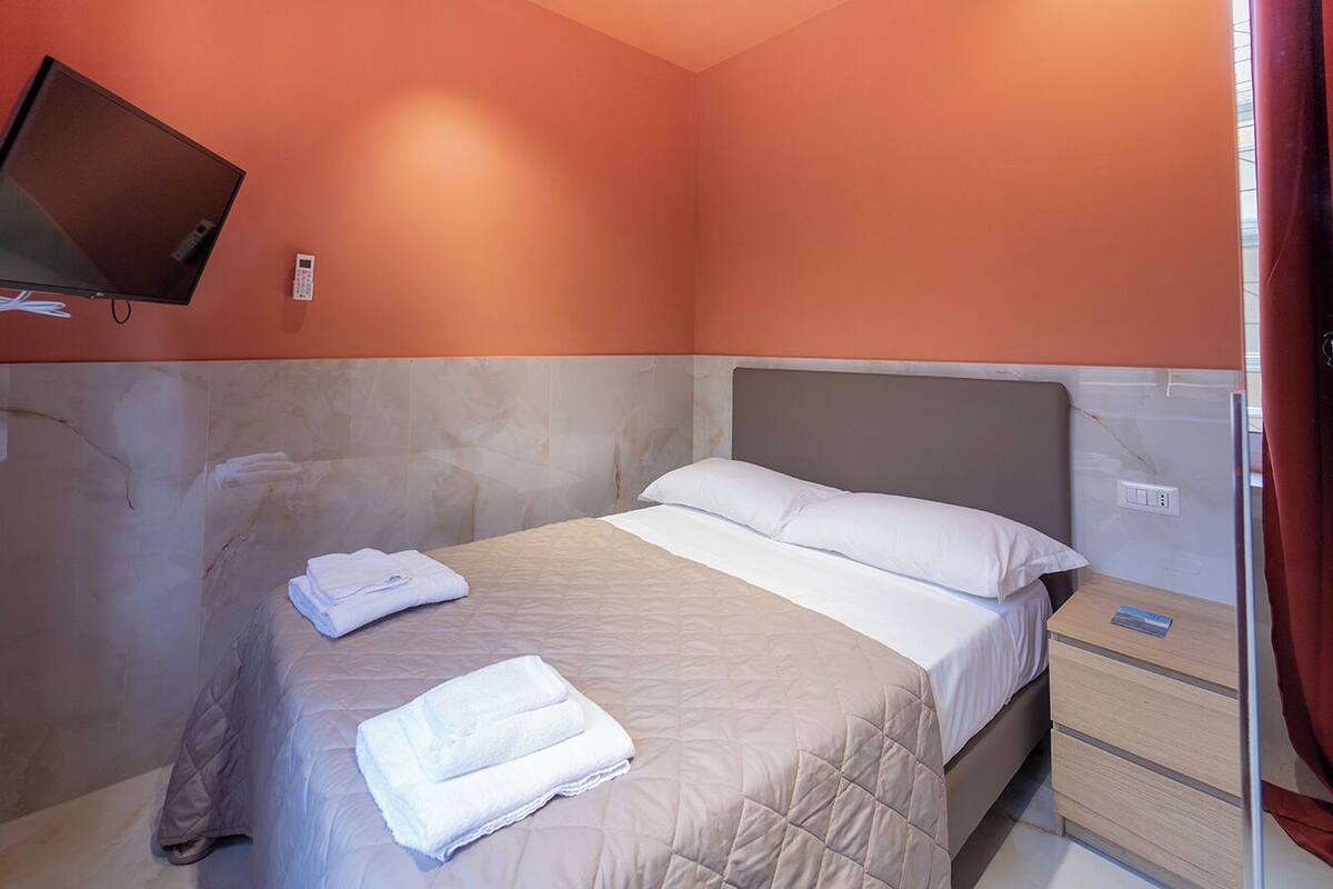 Een hotelkamer met een bed, een aan de muur bevestigde televisie, een nachtkastje en een stopcontact.