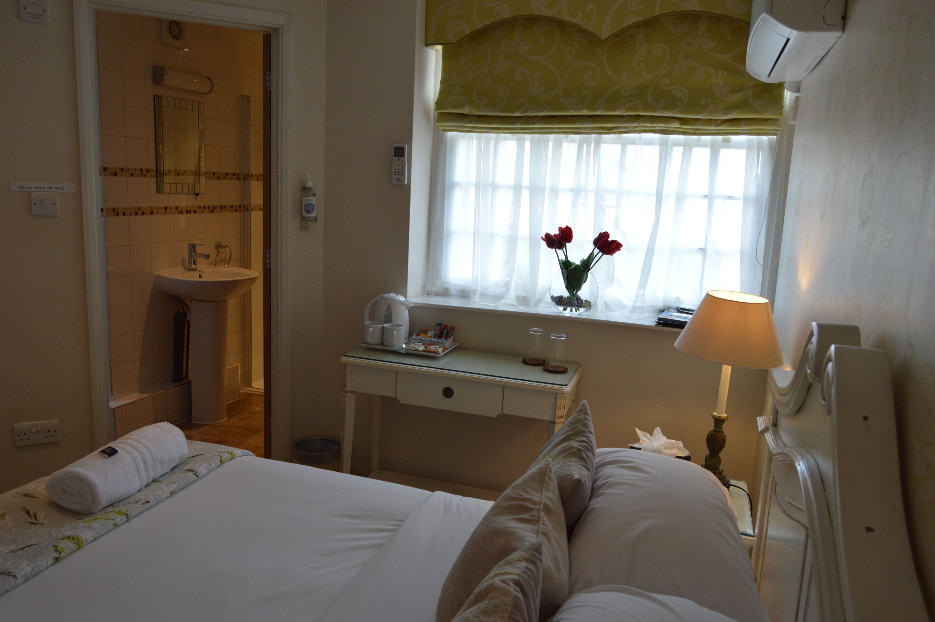 A hotel room with a bed, a bedside table with a lamp, a window with a view of the outside, and a bathroom with a sink and mirror.