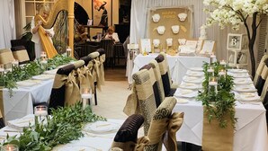 Un comedor formal con mesas dispuestas para una comida, destacando manteles blancos, cubiertos dorados y decoración con follaje verde.