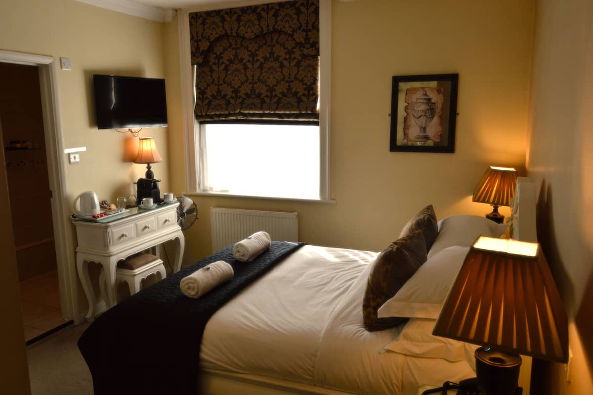 Habitación de hotel con cama, lámparas de noche, cómoda, televisor, ventana con cortinero y un cuadro en la pared.