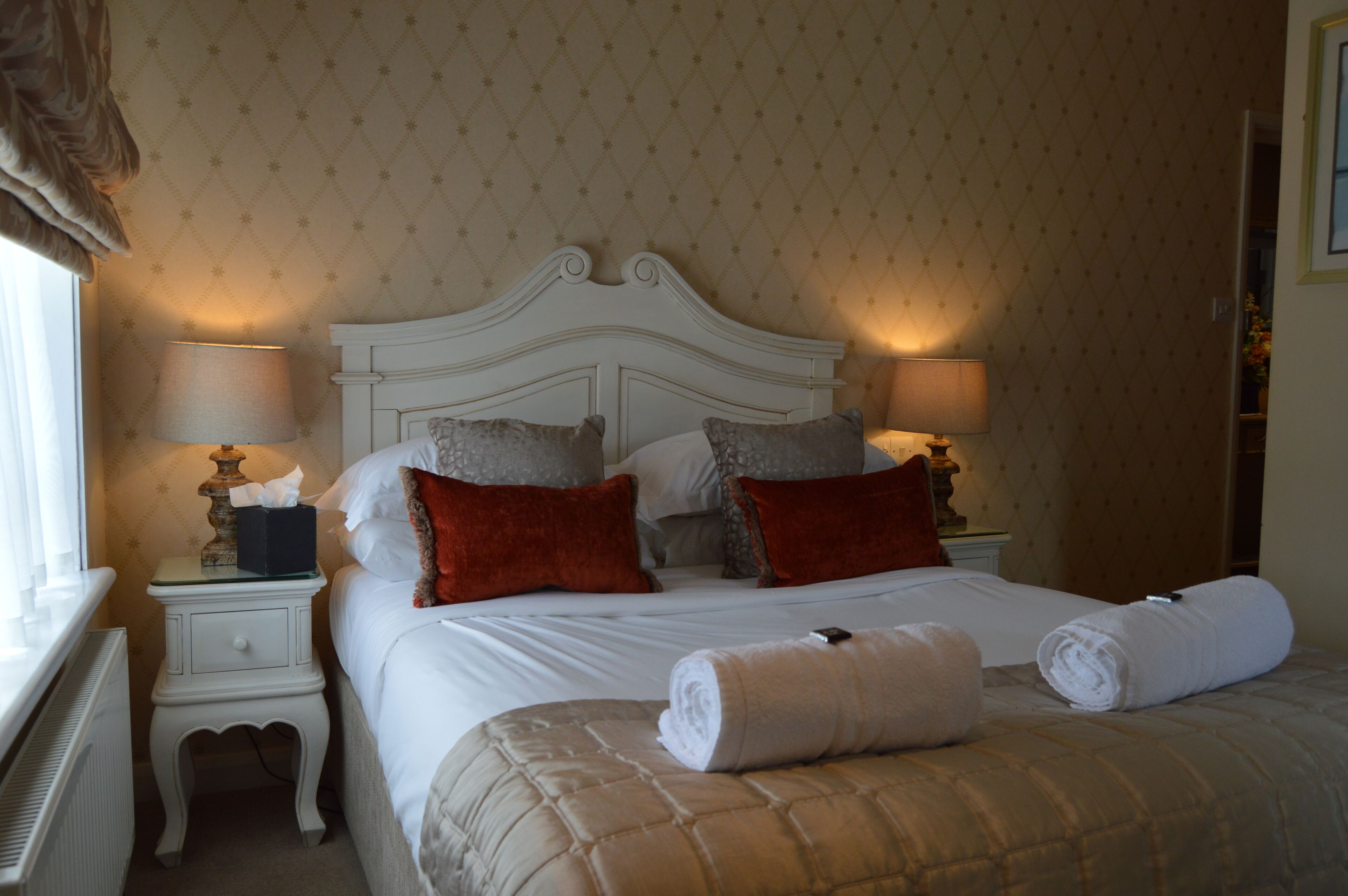 Un lit bien fait, avec des draps blancs, deux oreillers rouges et une couverture beige. Deux lampes de chevet sont placées de chaque côté du lit.