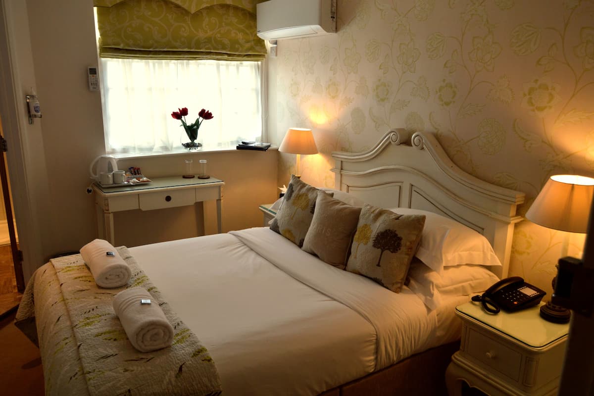 Una cama bien hecha con almohadas, una lámpara de noche, una mesita de noche con un teléfono y una ventana con vista a flores.