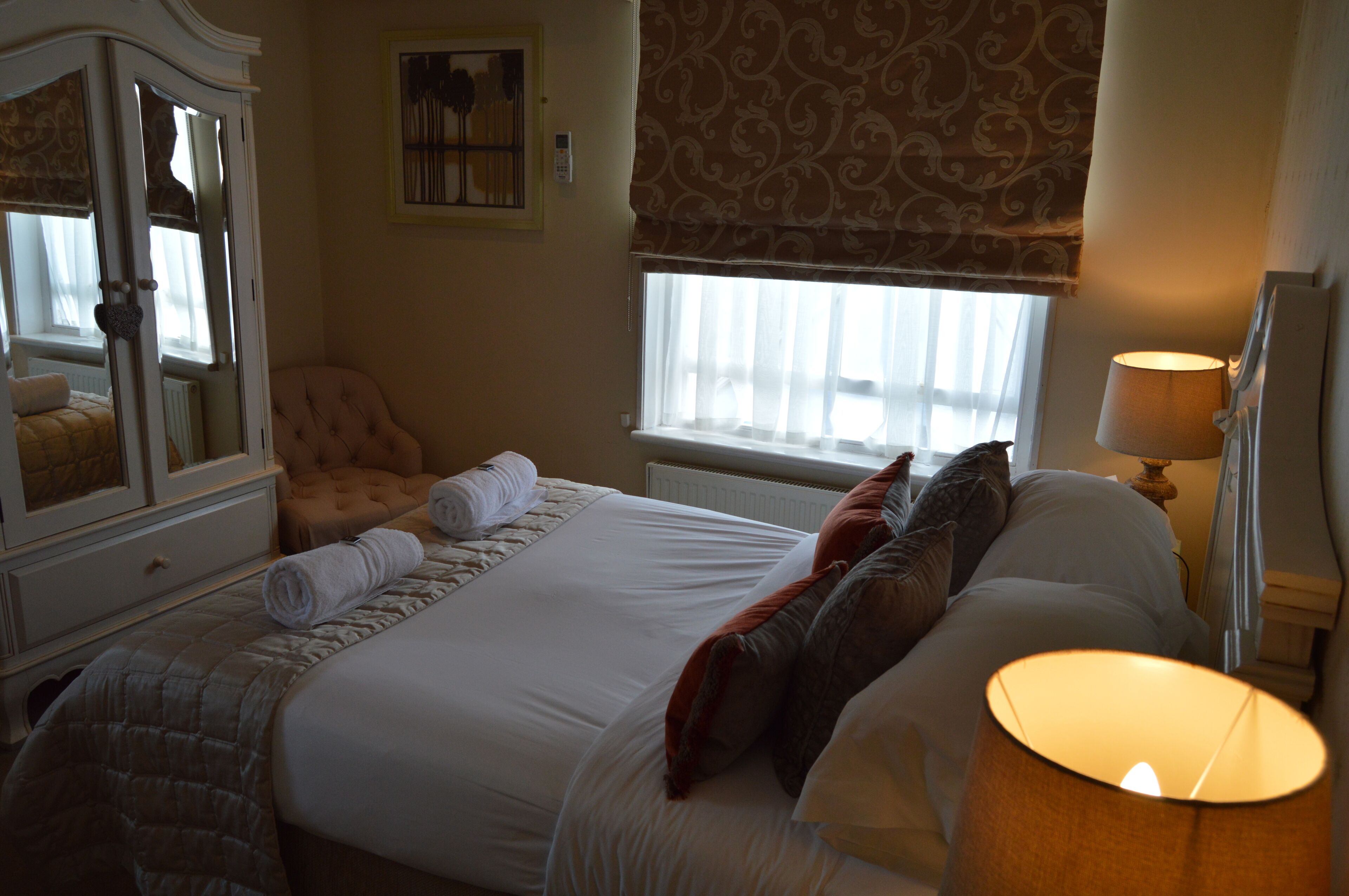 A neatly made bed with pillows and a lamp, a chair, and a dresser with a mirror.